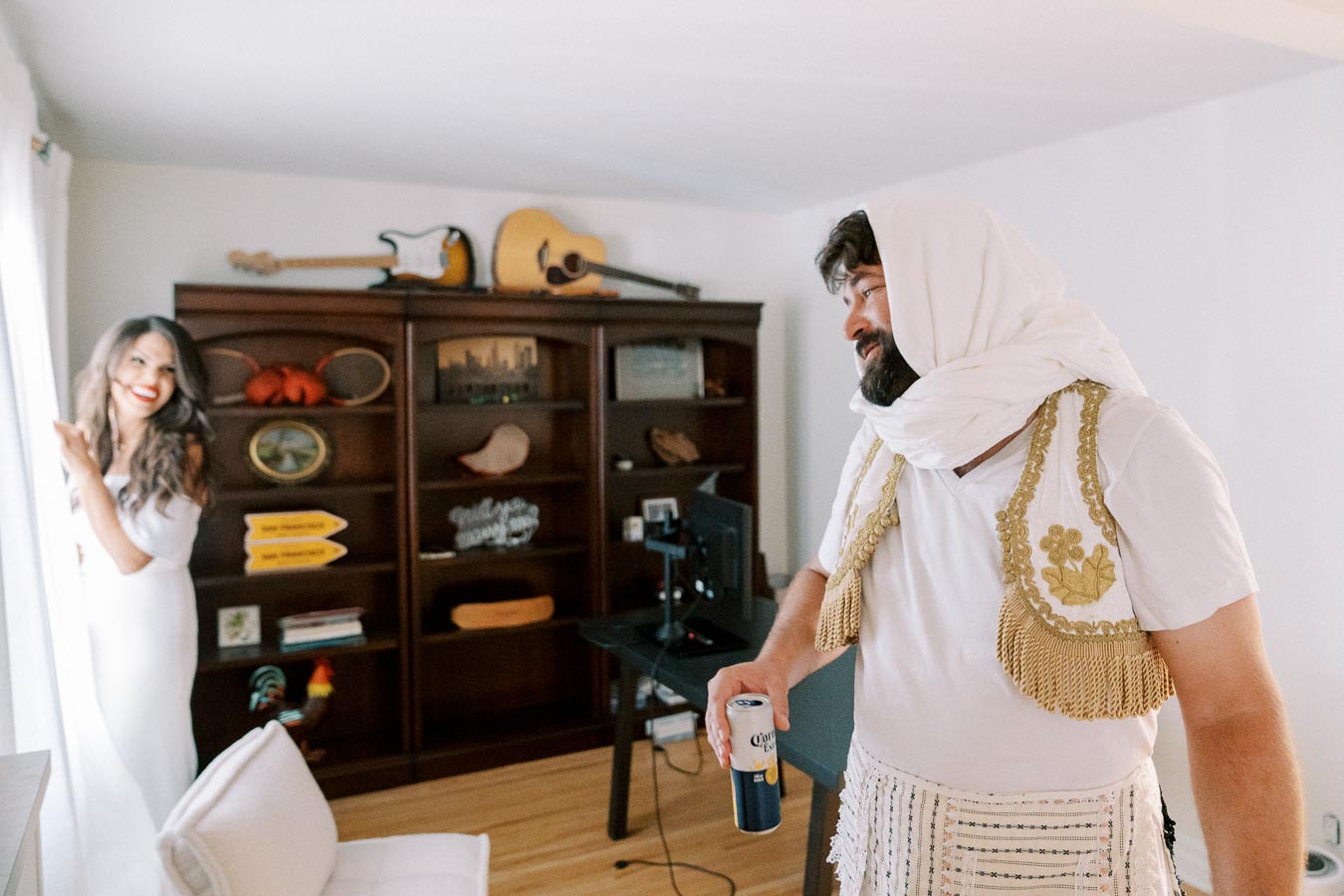 A man dressed in traditional attire holding a beverage can stands in a living room, smiling towards a woman in a white dress near a window, with a bookcase filled with decorative items in the background.