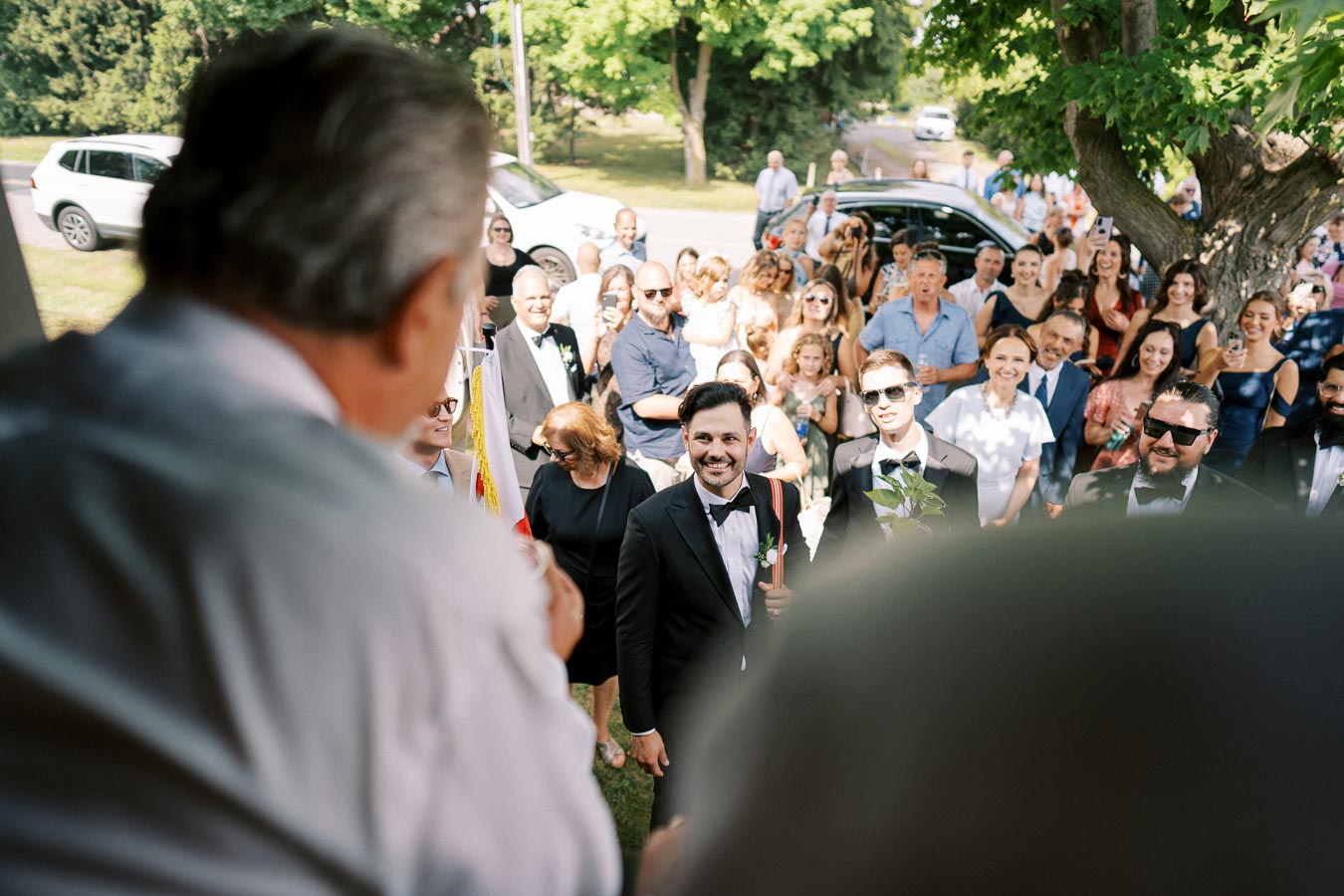 A groom in a tuxedo smiling at a wedding ceremony, surrounded by a large group of guests outdoors, with trees and cars in the background.