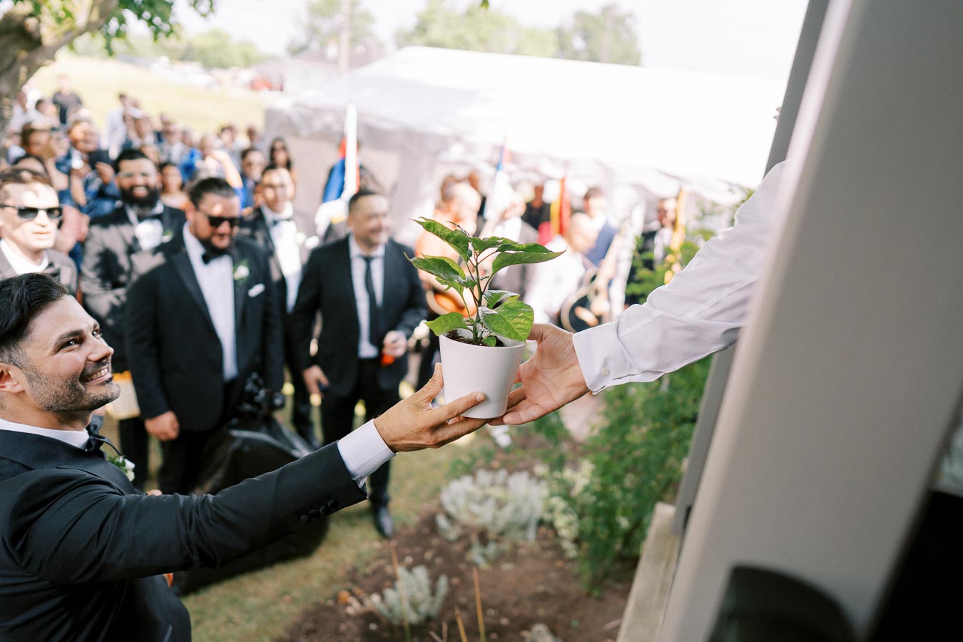 A man smiles while receiving a potted plant as a gift during an outdoor formal gathering, with a crowd of well-dressed guests in the background.