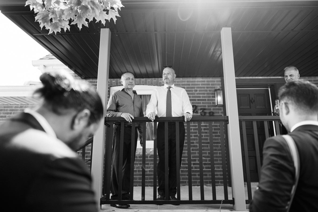 Black and white photo of two men standing on a porch, surrounded by others. They appear to be engaged in a conversation or speech, with a brick building in the background and sunlight filtering through tree leaves overhead.