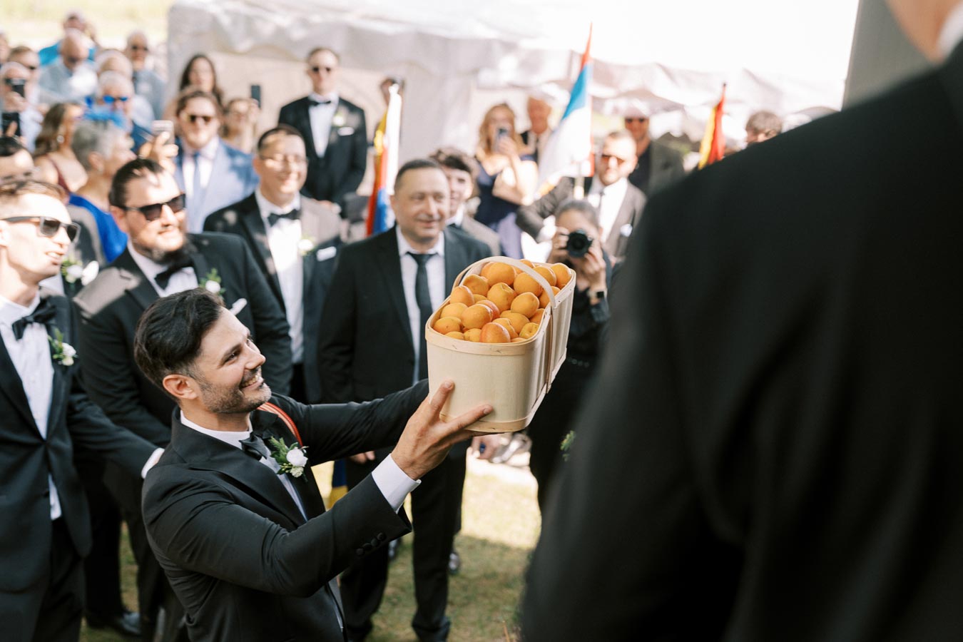 A man in a black tuxedo presents a basket of apricots to a group of people dressed formally at an outdoor event.