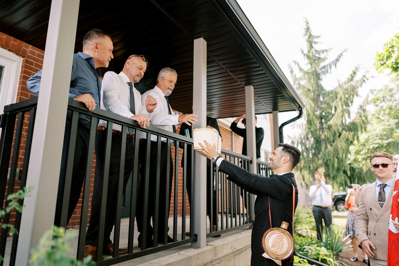 A group of men in formal attire stand on a porch, engaging with another man holding a basket, set outdoors with trees and a brick building in the background.