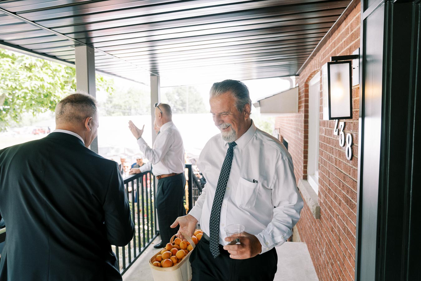 A man in a white shirt and tie smiling while holding a basket of apricots and a glass on a porch, with two men in the background engaged in conversation. The setting is bright, with natural light and a brick wall nearby.