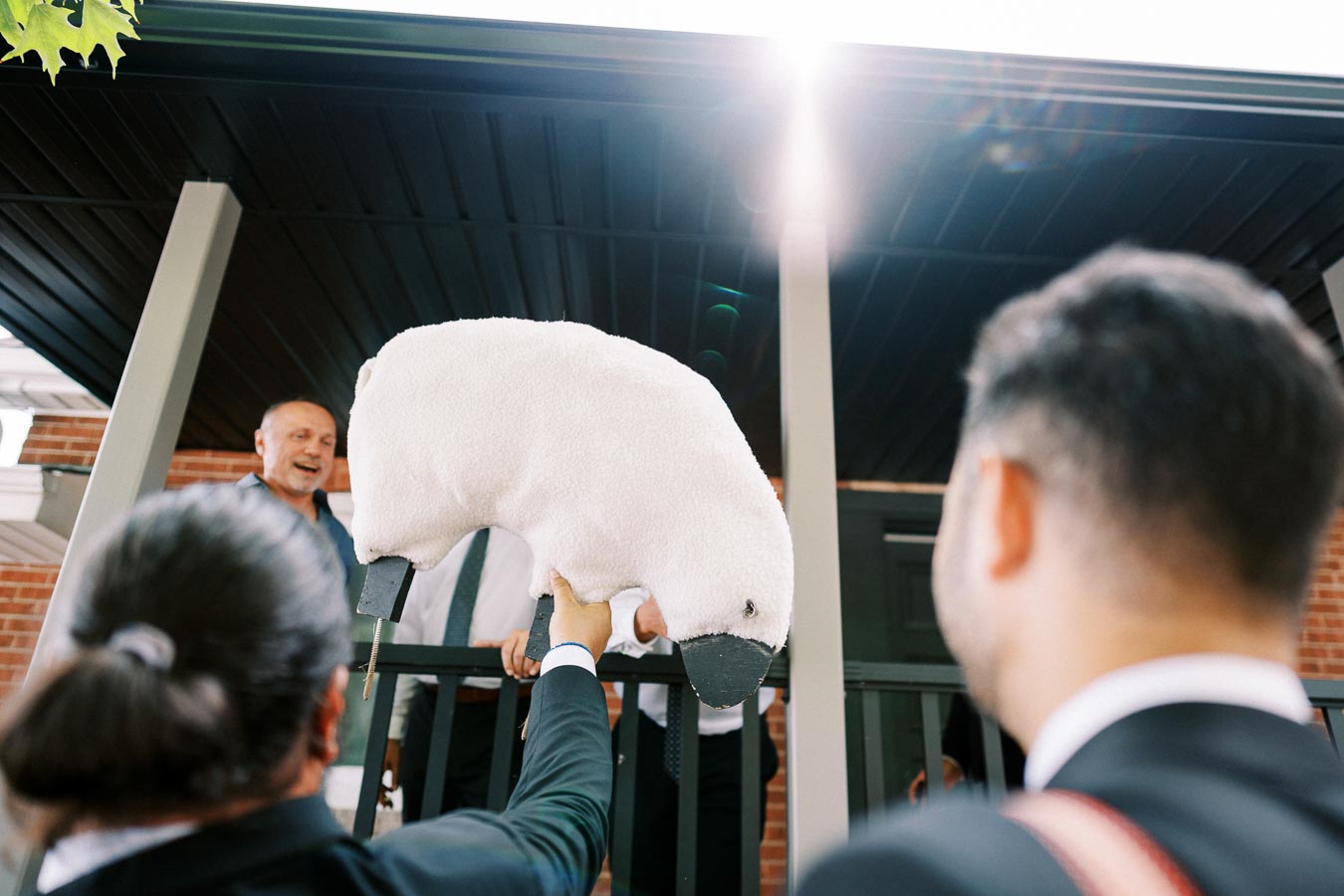 A person in a suit holding up a plush sheep toy at a porch, where another person is smiling, with sunlight shining in the background.