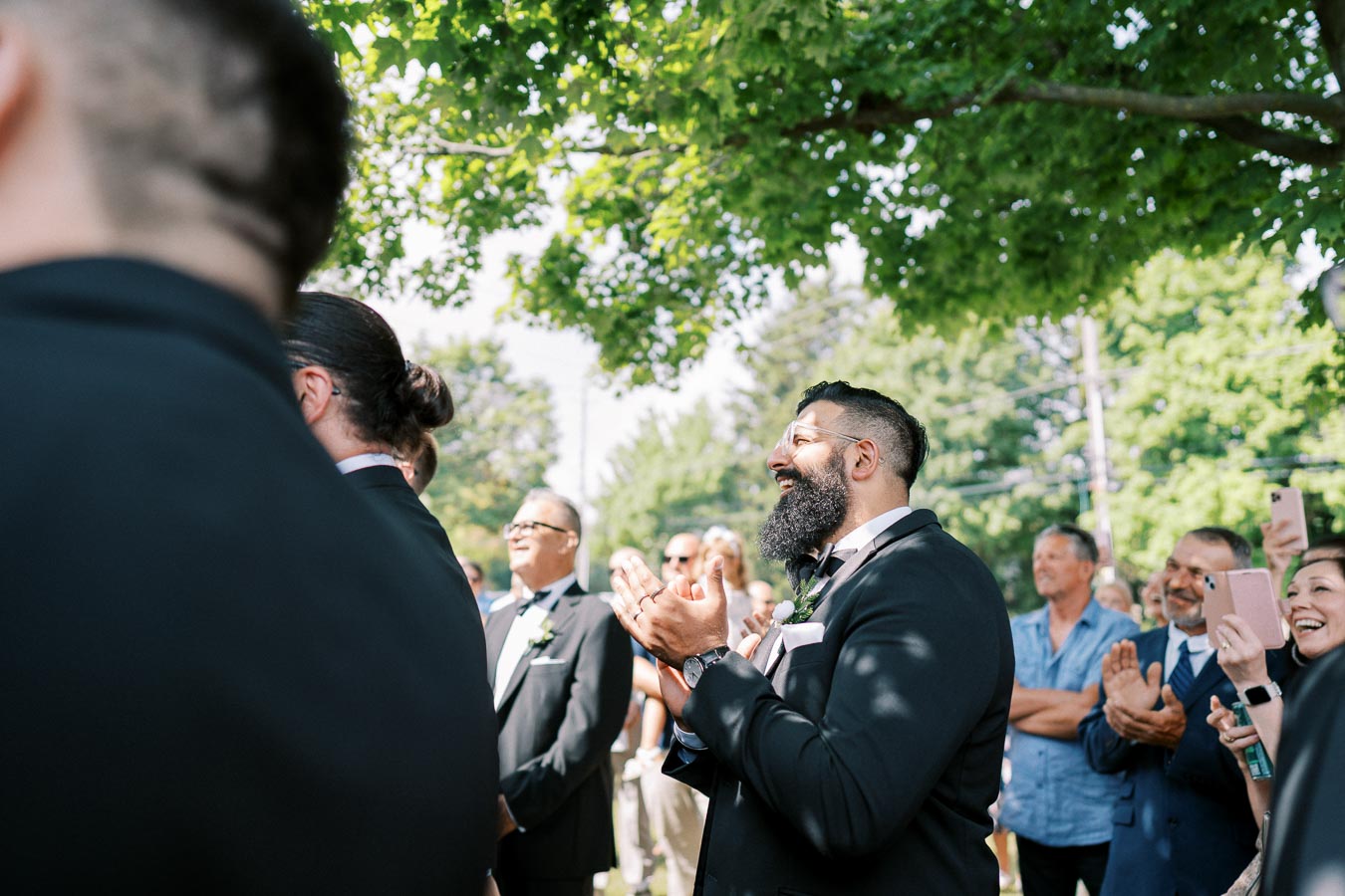 Group of people clapping and smiling at an outdoor event, dressed in formal attire under a leafy tree.