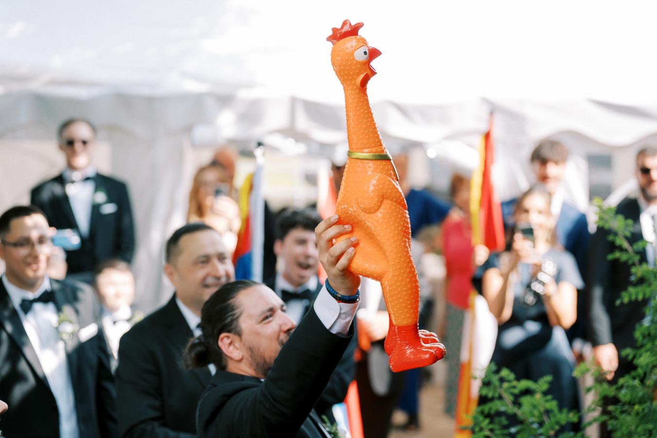 A man in a suit holds up a large rubber chicken enthusiastically at an outdoor event, surrounded by smiling guests in formal attire.