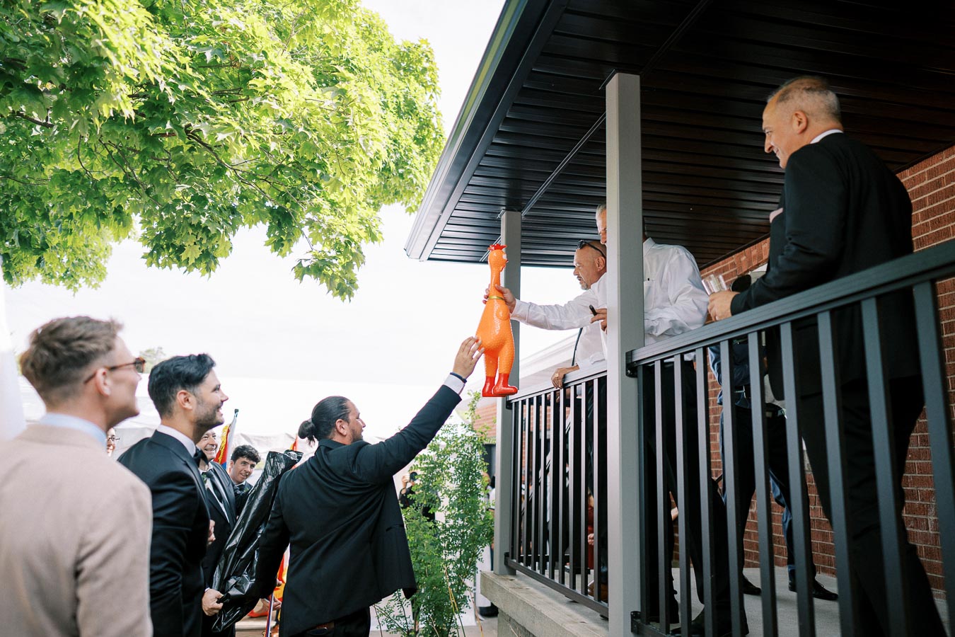 A group of men in suits and dress shirts stand on a porch and exchange a large, inflatable rubber chicken. One man reaches from the ground while another leans over the railing. The scene is set against a backdrop of lush green trees and a brick building.