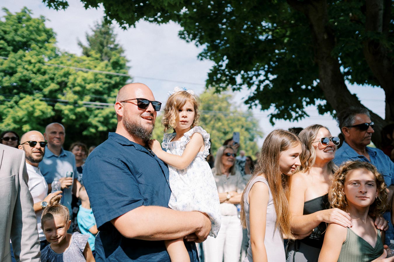 A joyful gathering of people outdoors under trees, featuring a bald man in sunglasses holding a young girl, surrounded by smiling individuals enjoying a sunny day.