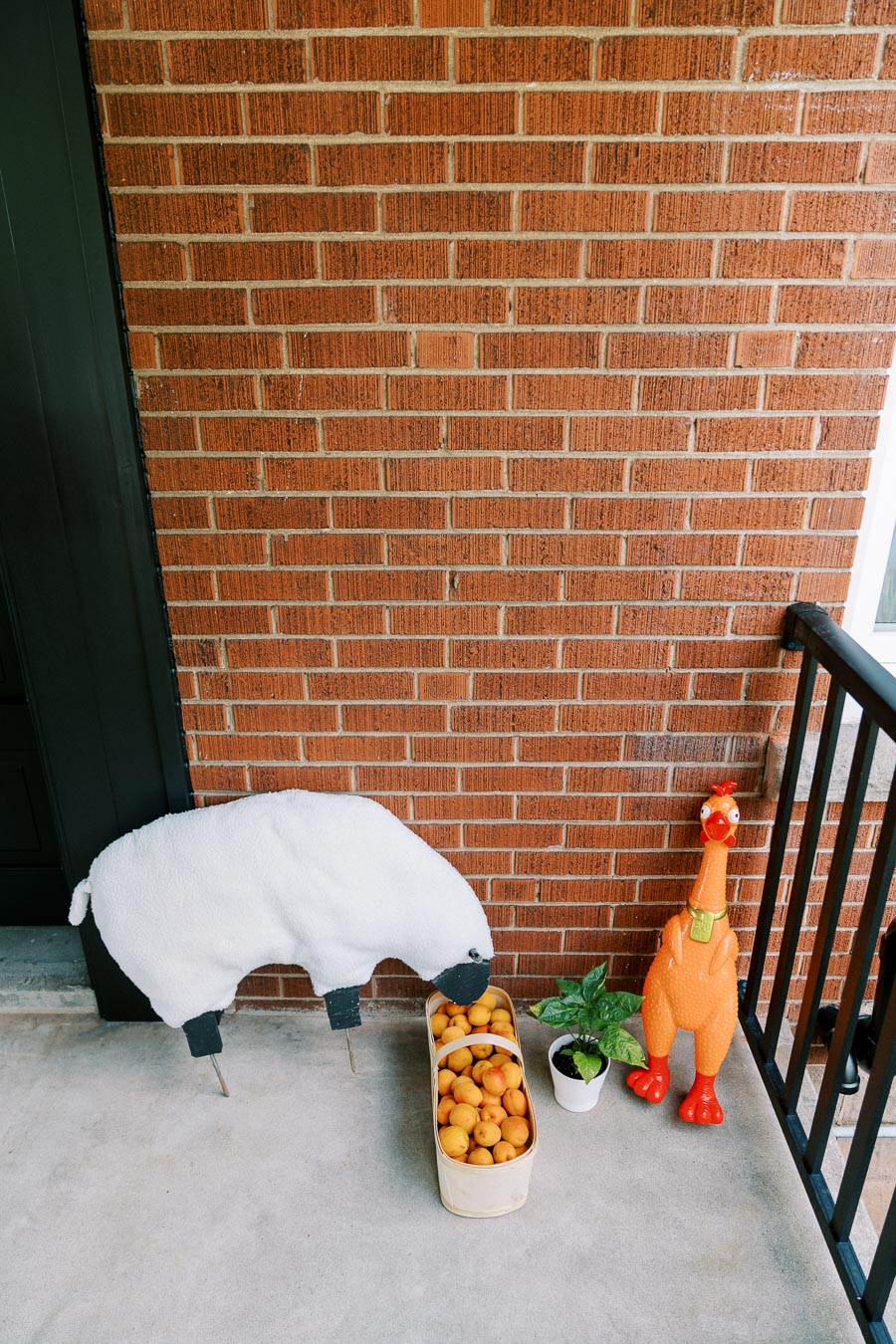 Quirky porch scene with a plush sheep sculpture, a basket filled with apricots, a small potted plant, and a large rubber chicken statue, set against a red brick wall background.