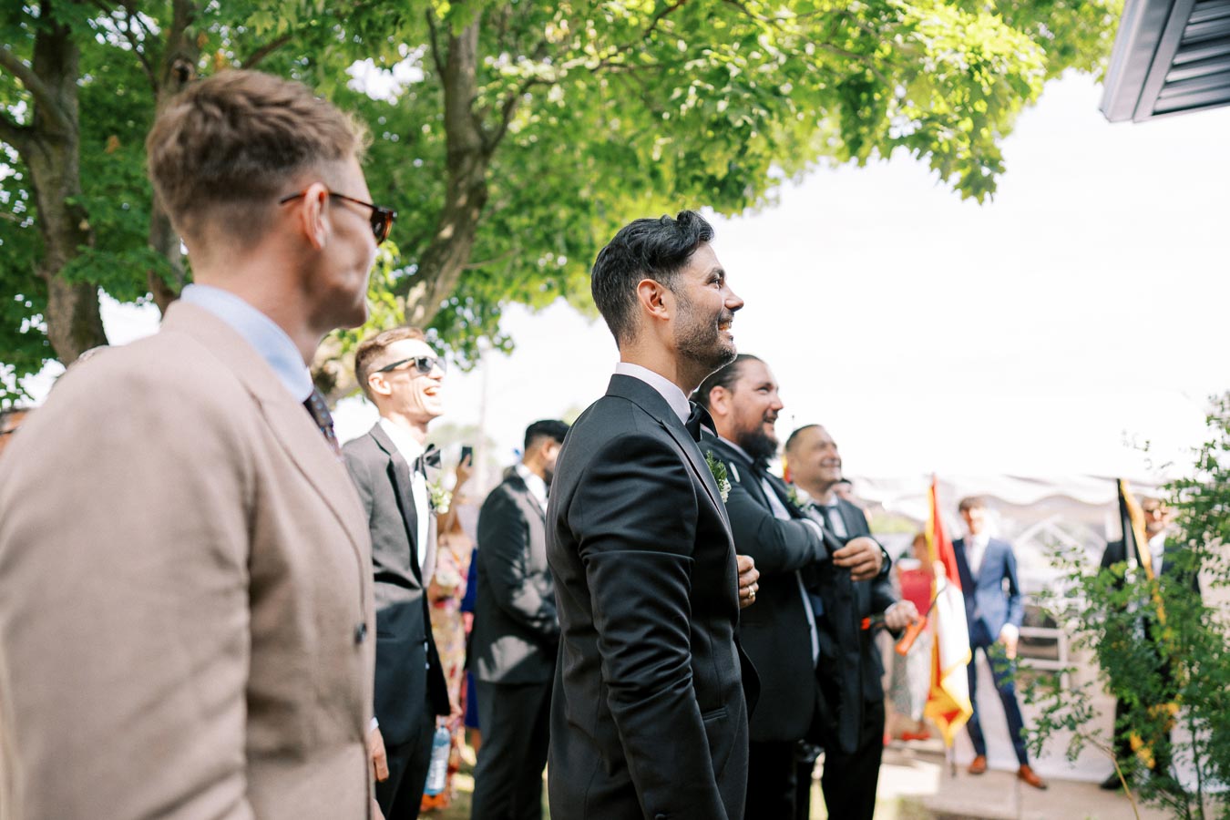 Group of well-dressed groomsmen laughing and smiling outdoors at a wedding ceremony, surrounded by greenery and sunlight.
