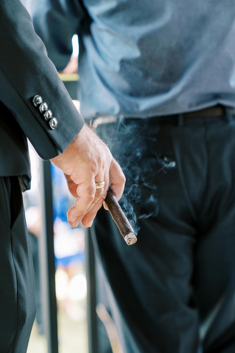 A person in a suit holding a lit cigar, emitting smoke, during a social event.