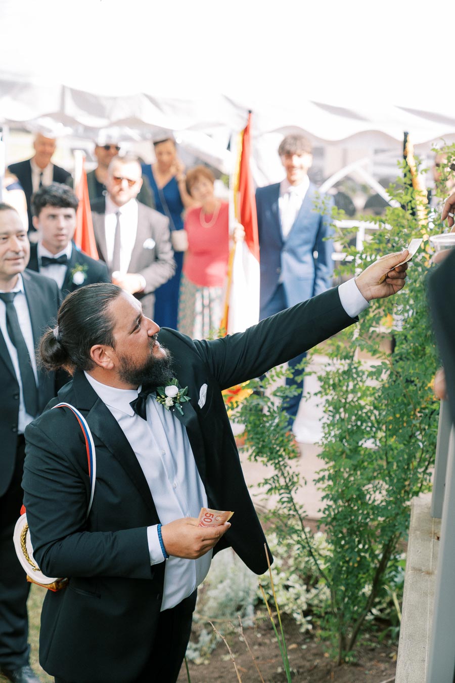 A formally dressed man with a beard offers a note during an outdoor ceremony, surrounded by a group of people in elegant attire, underneath a white canopy.