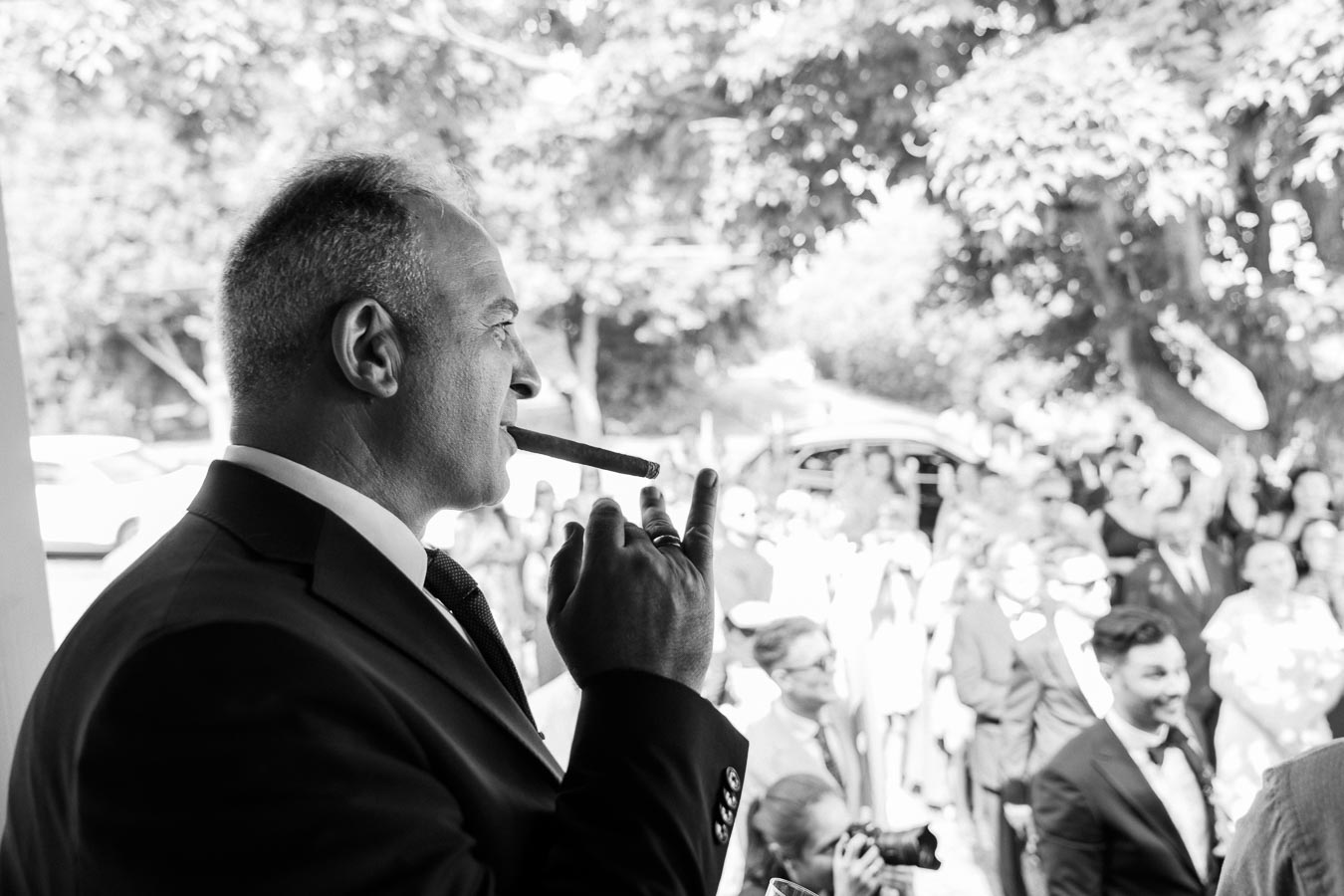 Black and white image of a distinguished man in a suit enjoying a cigar at an outdoor event, surrounded by a crowd of people in formal attire amidst a backdrop of trees.