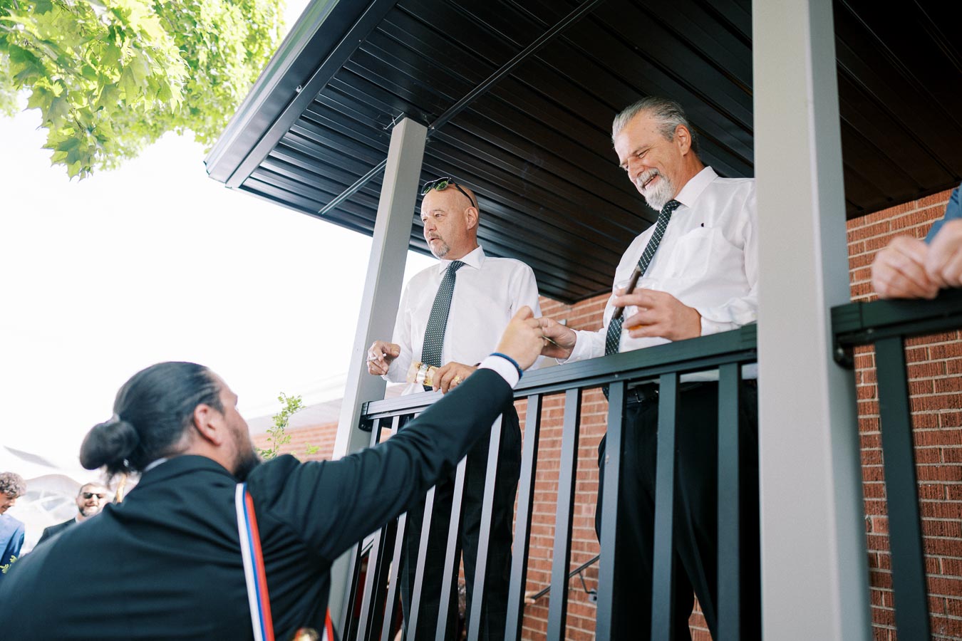 A group of men in formal attire socializing on a covered porch, with two men standing and holding cigars and drinks, and another man with a beard facing them, possibly engaged in conversation. The setting includes a black railing and brick walls, suggesting an outdoor gathering or event.