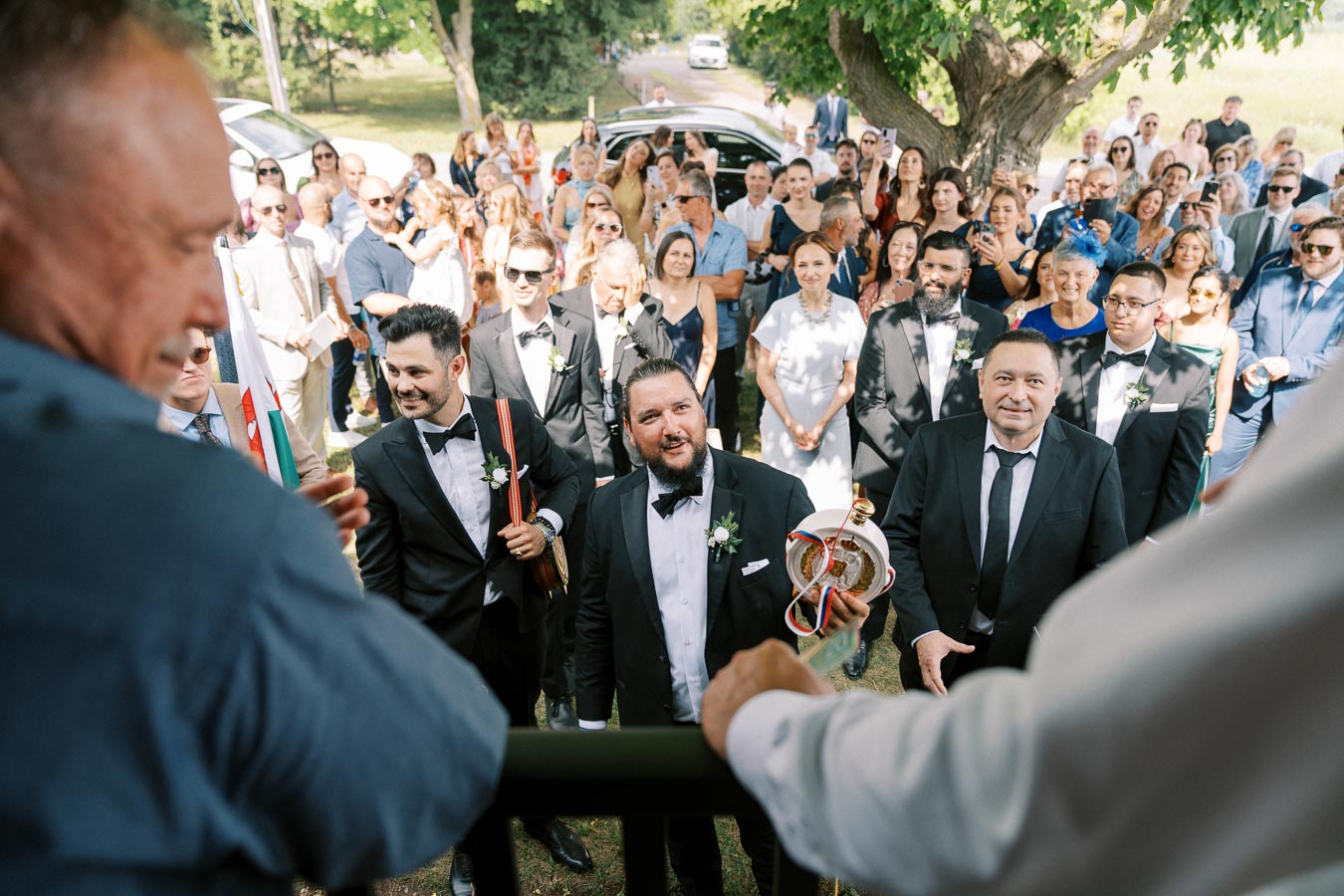 Groom and guests gather outdoors for a wedding ceremony, with the groom holding a traditional decorative item, surrounded by smiling attendees in formal attire under a lush green tree.