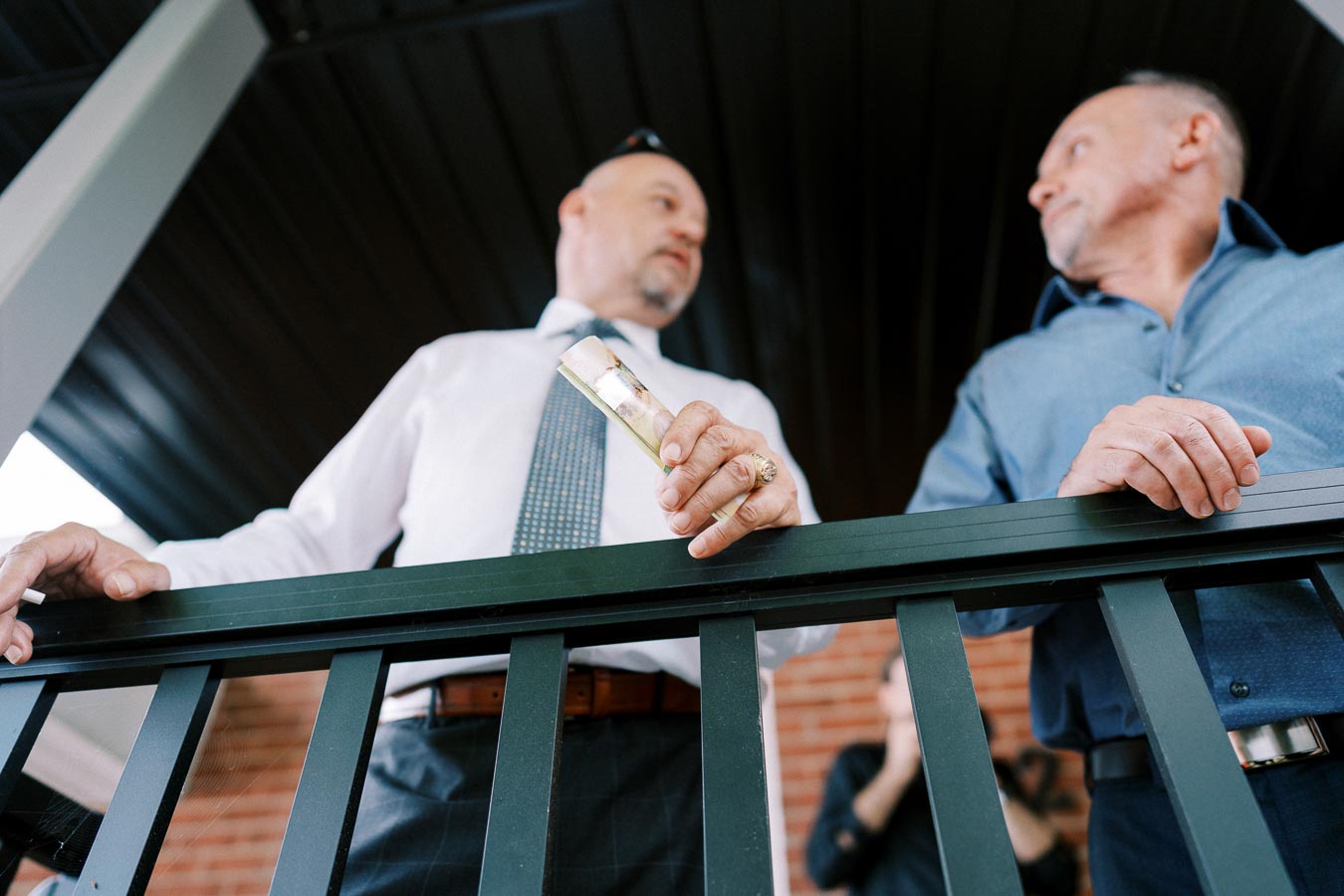 Two men in formal attire standing on a balcony, one holding a champagne glass, engaged in conversation.