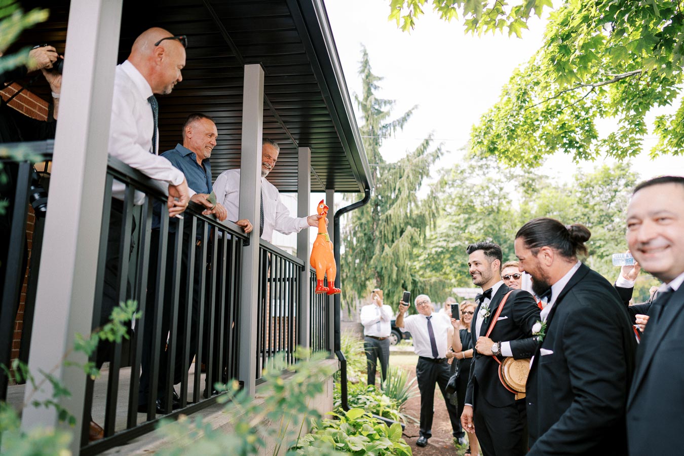 A group of well-dressed men, one holding a novelty rubber chicken, enjoy a humorous moment on a porch at an outdoor event, surrounded by trees and greenery.