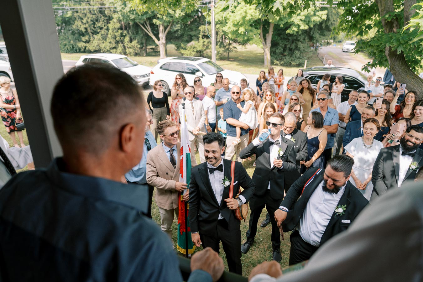A large group of people gathered outdoors for a wedding celebration, with guests in suits and dresses, enjoying a sunny day.