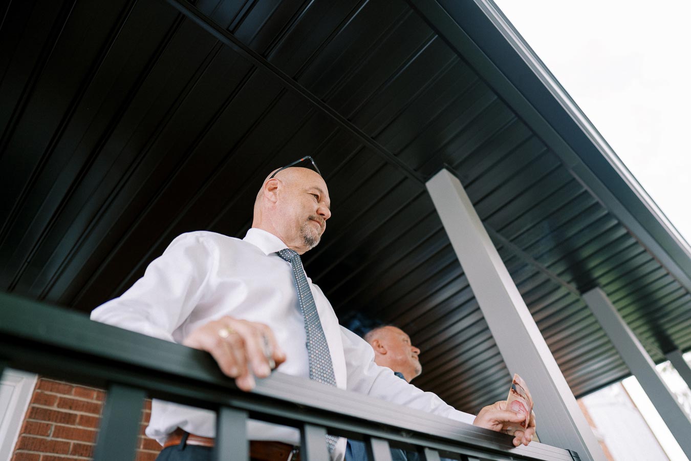 Two men in white dress shirts and ties stand on a balcony under a dark roof, gazing into the distance. The perspective from below highlights the architectural design of the building they are on.