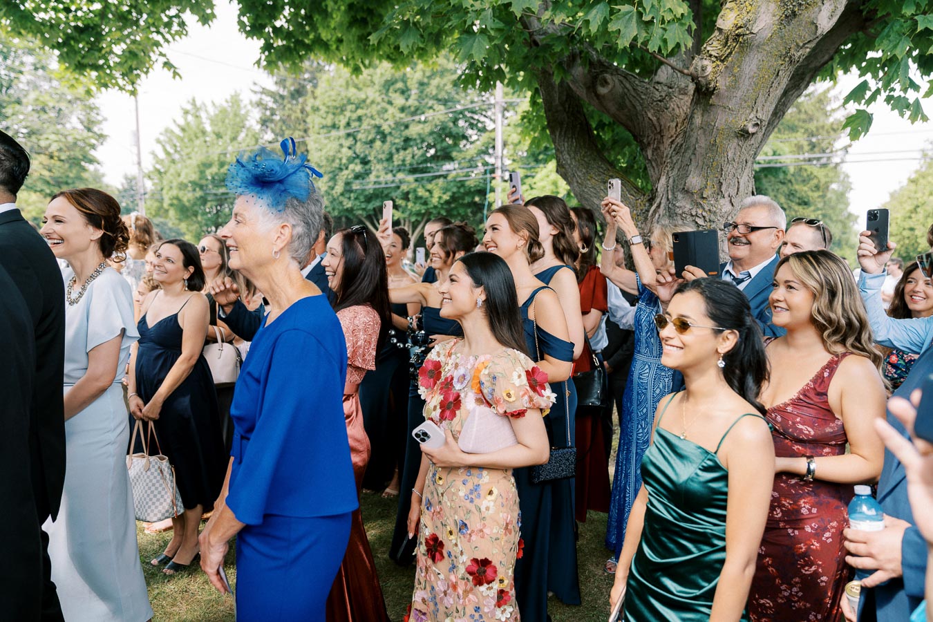 A joyful crowd at an outdoor wedding, featuring guests in colorful dresses and suits, smiling and capturing the moment with their smartphones under a large tree.