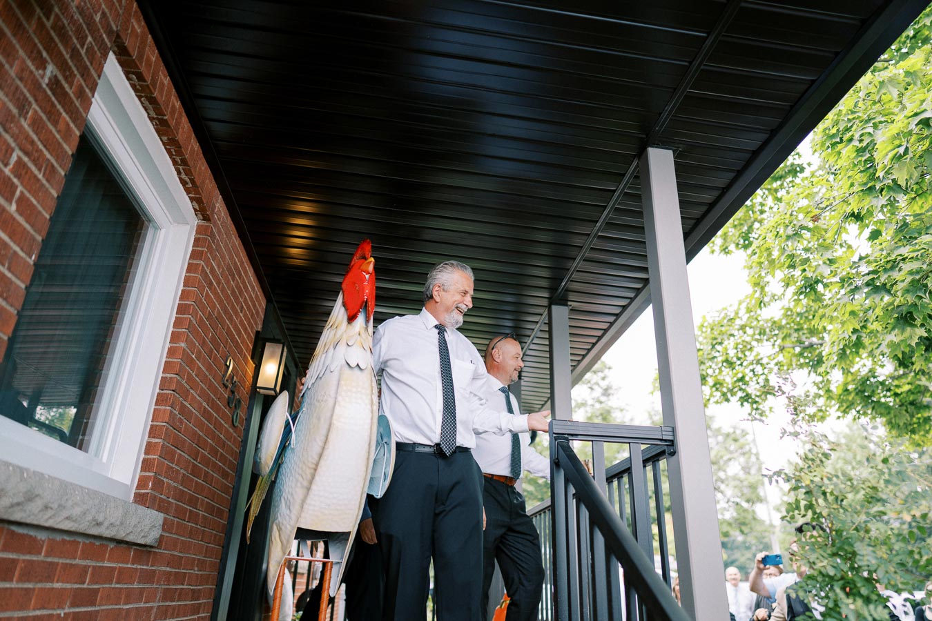 Two men in formal attire carrying a large decorative rooster under a porch, with a brick building and green trees in the background.