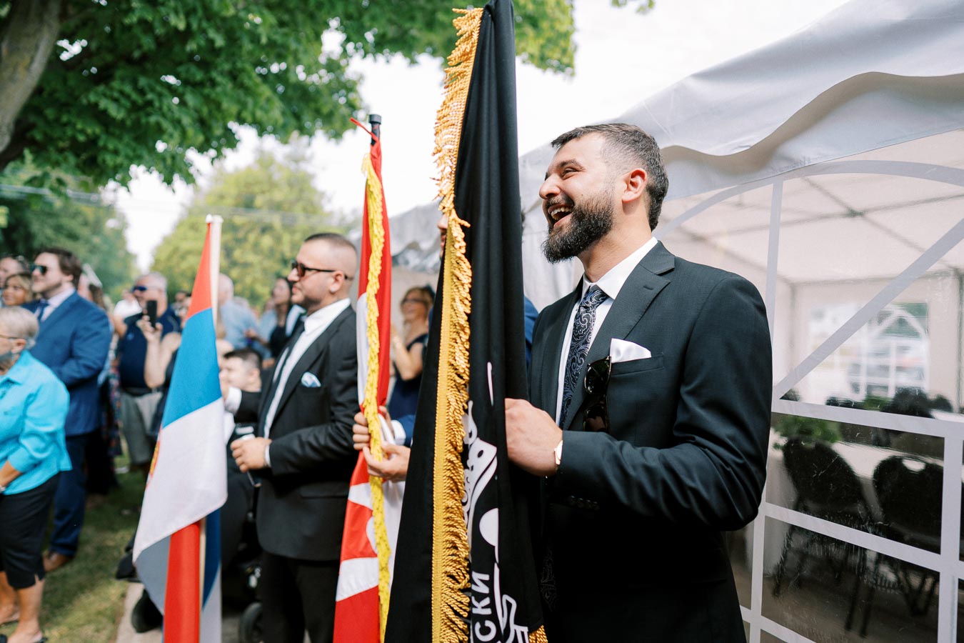 A man in a suit holds a ceremonial flag at an outdoor event, surrounded by people dressed formally under a tent, with trees and clear skies in the background.