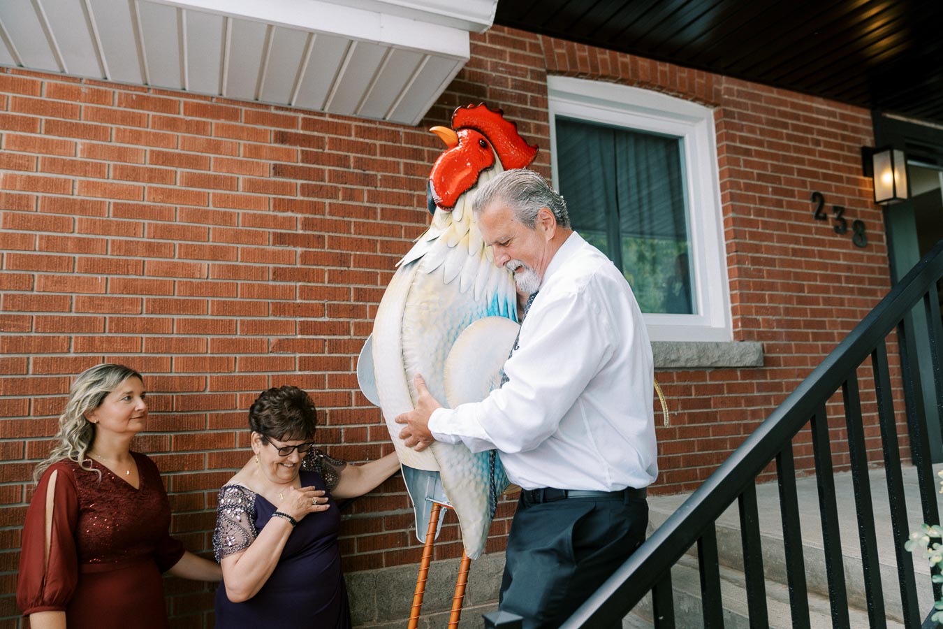 Senior man playfully embraces large decorative rooster statue, two women smile nearby on brick house porch.