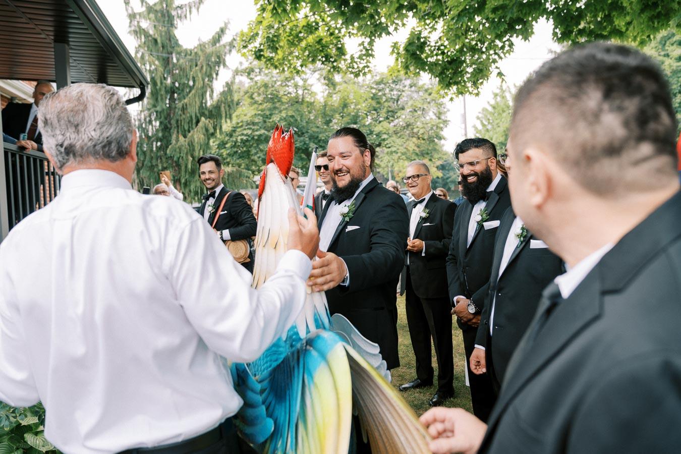 A group of well-dressed men gathered outdoors engaging in a joyful celebration involving a large, colorful rooster sculpture. The scene is lively, set against a backdrop of greenery and blue skies, capturing a moment of cultural or festive significance.