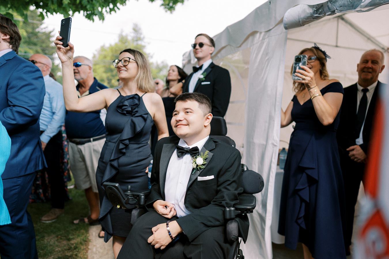 Group of people attending an outdoor formal event, with a smiling person in a wheelchair in the foreground wearing a tuxedo, surrounded by others capturing the moment with smartphones and cameras under a white canopy.