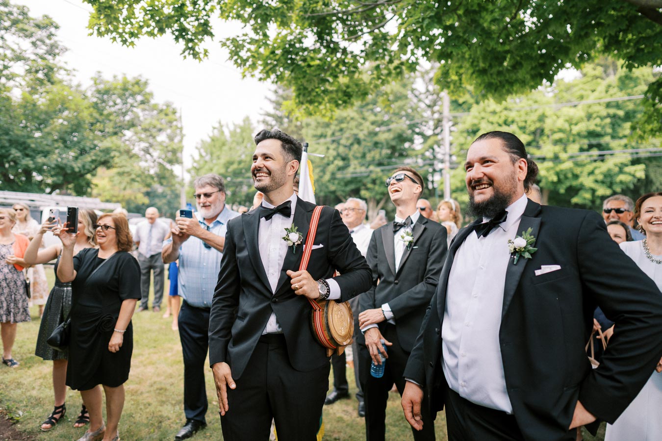 A group of men in formal suits and bow ties, smiling and standing outdoors during a celebration, surrounded by a crowd of people taking photos under green trees.
