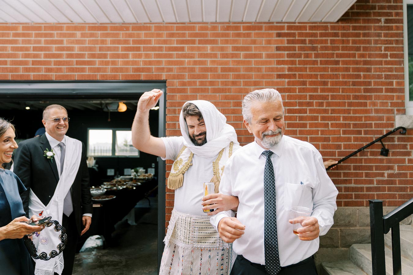 Group of people celebrating outdoors, with one man in traditional Albanian attire dancing joyously. Smiling guests are dressed in formal clothing, standing near a brick building entrance. Warm, festive atmosphere at a social gathering.