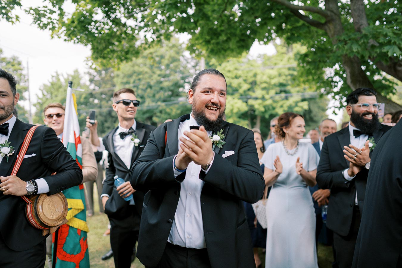 A joyful group of people clapping and smiling at an outdoor event, dressed in formal attire beneath leafy trees on a sunny day.
