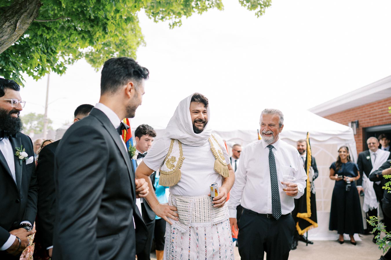 A joyful gathering of people at an outdoor event, with a man dressed in traditional attire and surrounded by others in formal wear, capturing a moment of celebration under a tree with a clear sky in the background.
