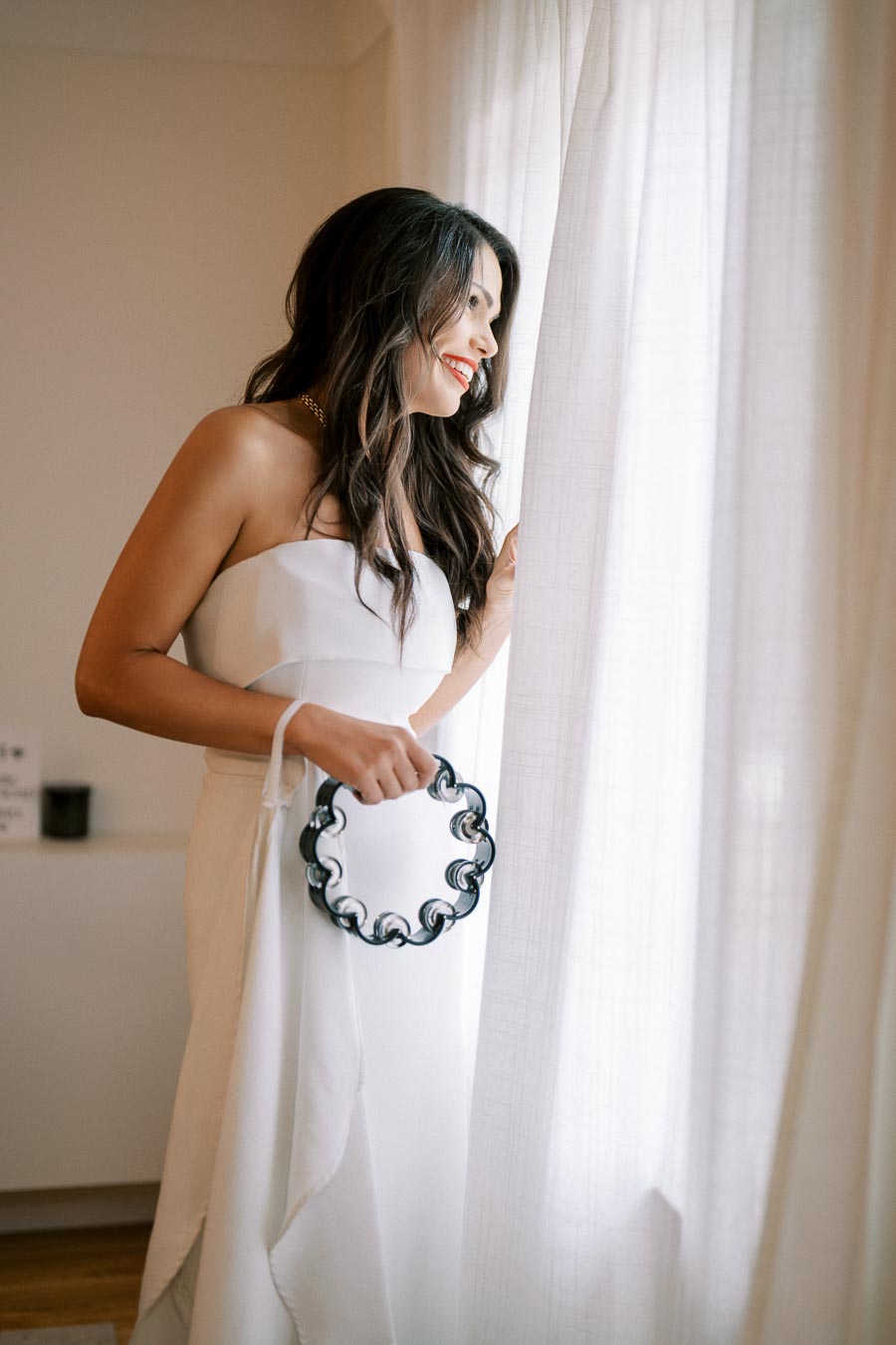 A woman in a white strapless dress stands by a window, holding a tambourine and smiling, with long hair cascading over her shoulders.