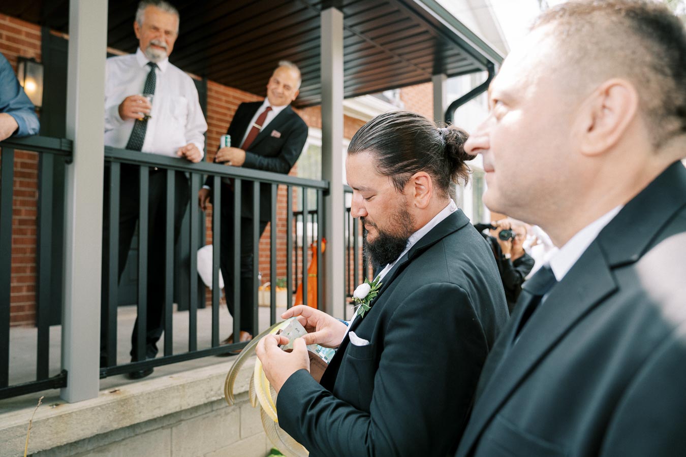 Groom preparing for wedding ceremony with groomsmen on a porch, showcasing formal attire and engagement in tradition.