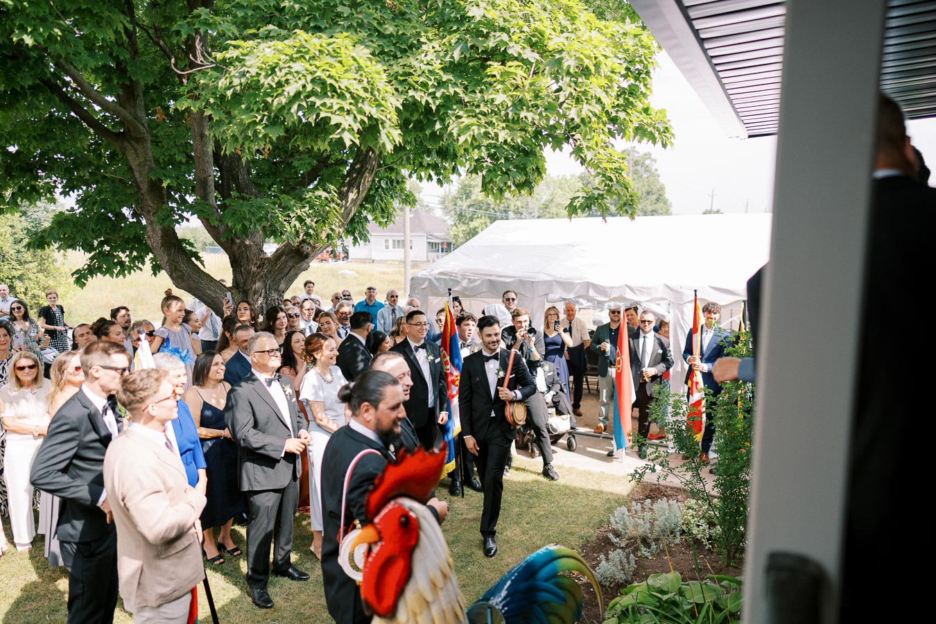 A large group of people dressed in formal attire gather outdoors under a tree for a celebration. A tent is set up in the background, and several attendees hold colorful flags. The event takes place on a sunny day, creating a cheerful atmosphere.