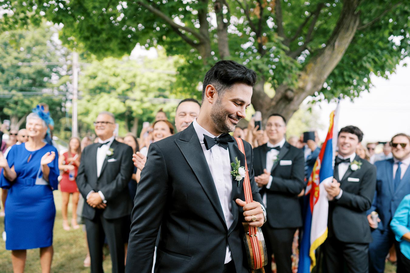 Groom in a black tuxedo smiling and holding a traditional stringed instrument, leads a cheering wedding crowd outdoors under a green tree.