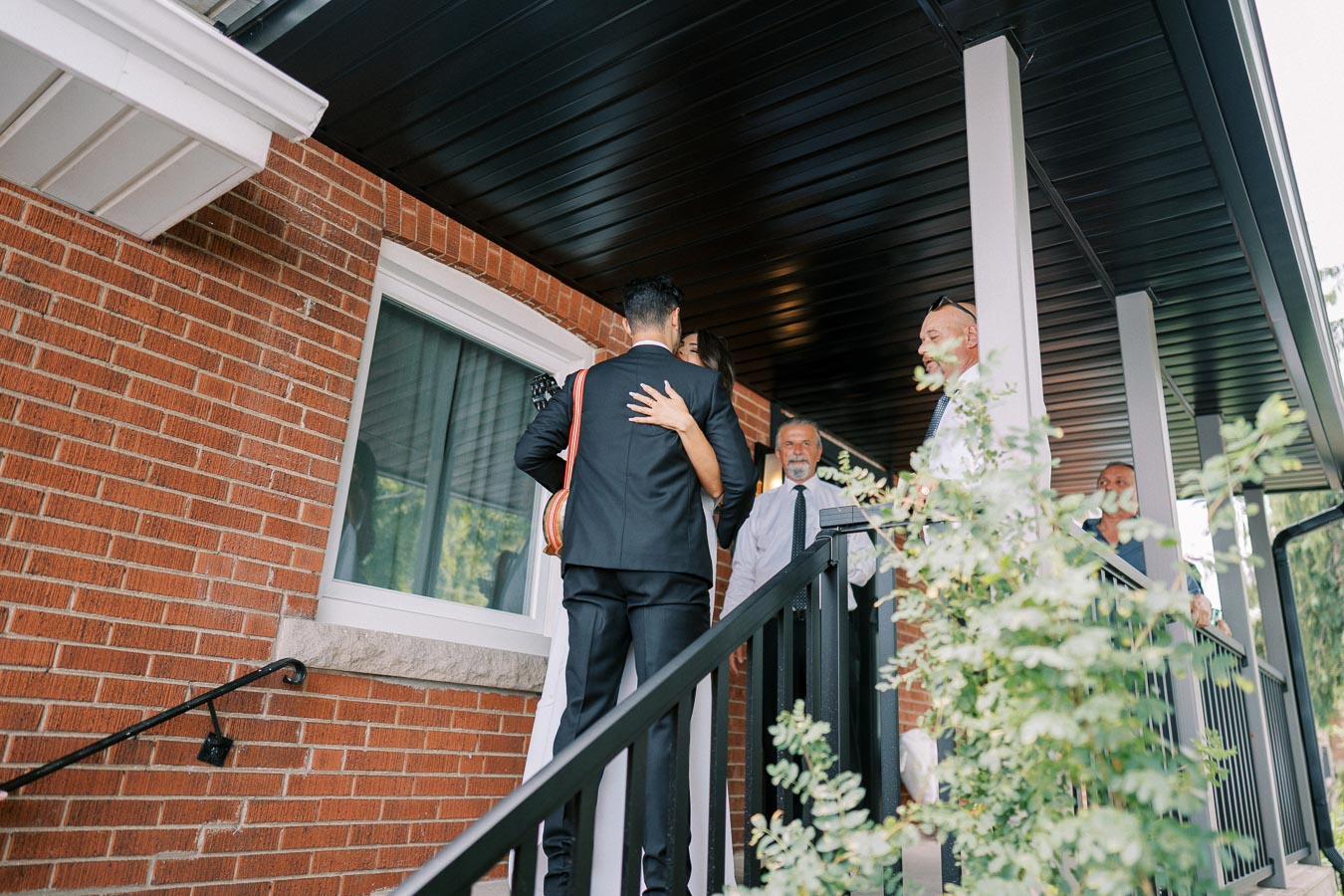 Group of people interacting on a house porch, including a man in a suit and a woman, with a brick exterior and black roof in the background