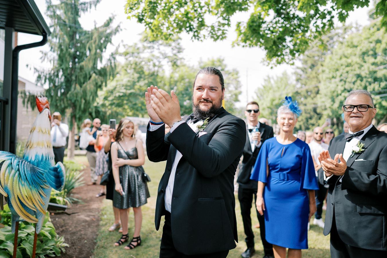 A group of wedding guests clapping outdoors, featuring a man in a black suit in the foreground, with a decorative rooster statue nearby and greenery in the background.