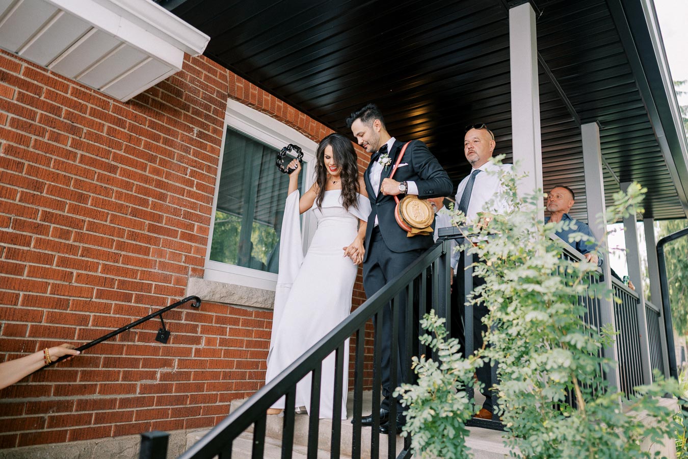 A joyful bride and groom in wedding attire descend stairs outside a brick building, with the bride holding a tambourine, accompanied by several wedding guests.