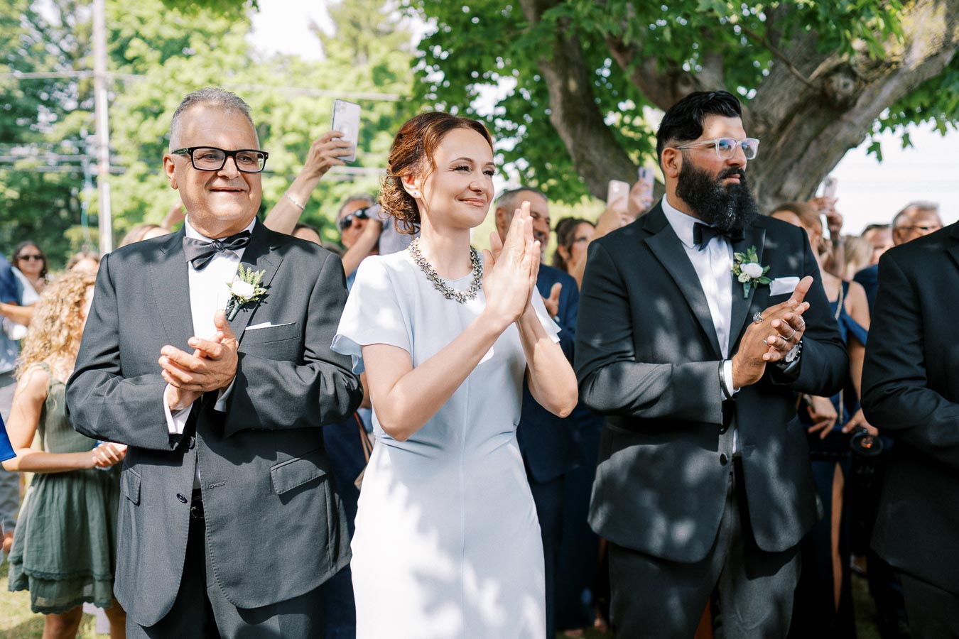 A group of elegantly dressed wedding guests, including a woman in a white dress and two men in black tuxedos, applauding outdoors under a tree, with a crowd and a background of greenery.