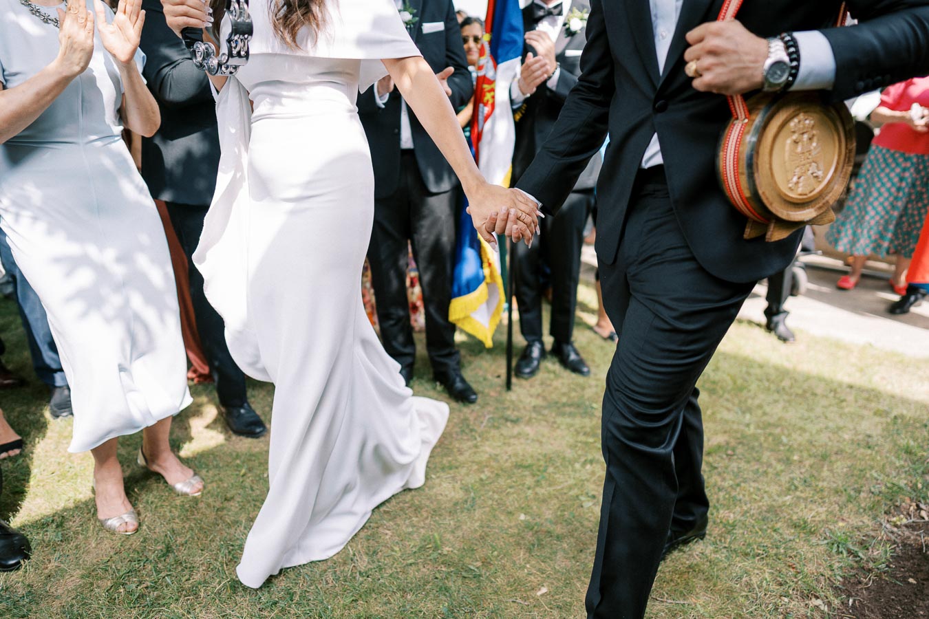 Elegant couple holding hands at a lively outdoor wedding celebration, wearing a white dress and black suit, surrounded by joyful guests.