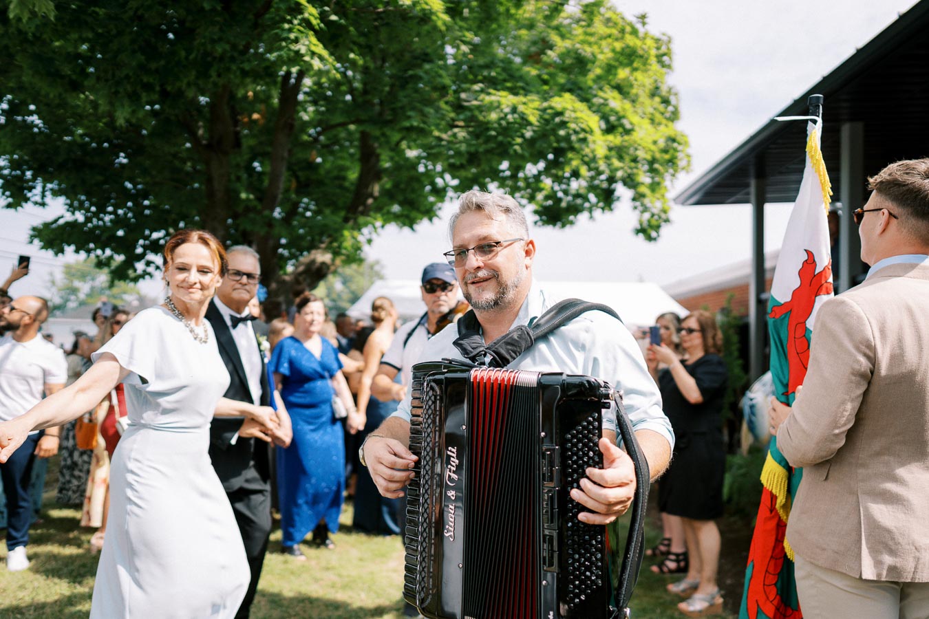 A lively outdoor celebration with a musician playing an accordion, surrounded by people dancing and enjoying the festive atmosphere under a clear sky.