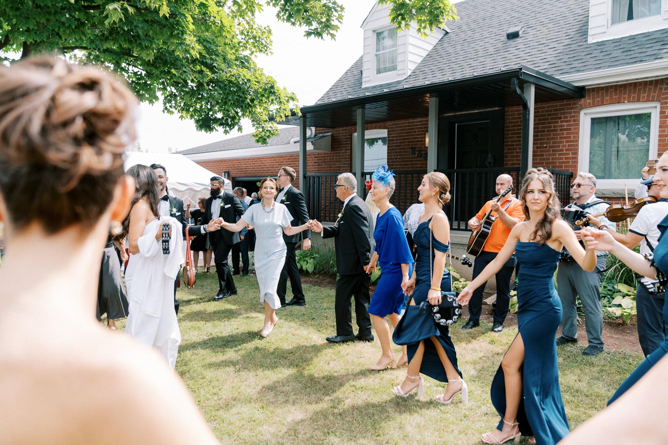 Outdoor wedding celebration with elegantly dressed guests dancing in a line, accompanied by live musicians playing guitars and violins, in a garden setting near a brick house under a large tree.