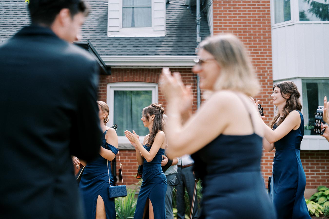 Group of women in elegant blue dresses clapping at an outdoor event, with a brick house in the background.