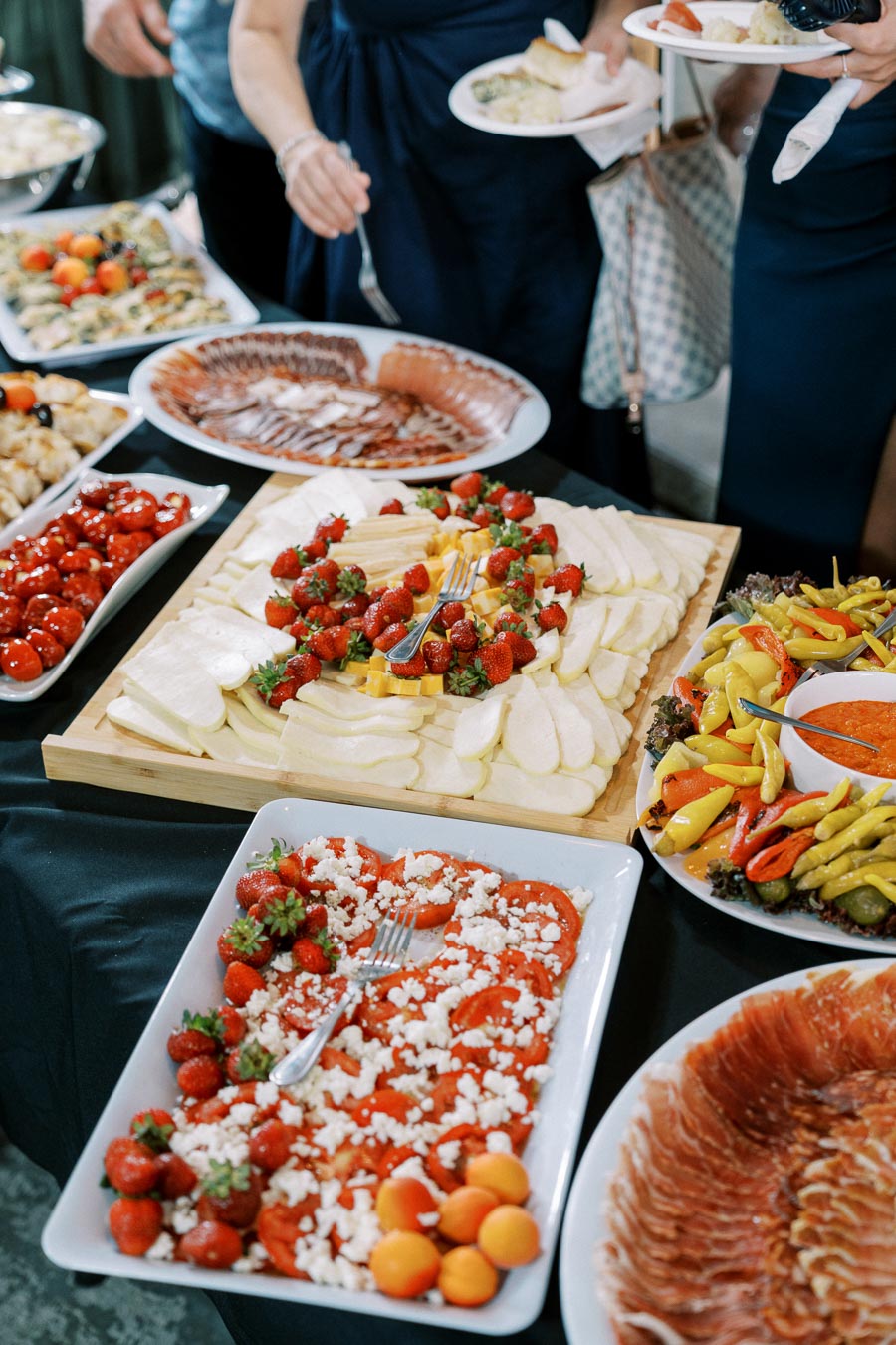 Buffet table with assorted appetizers including sliced cheeses, strawberries, tomatoes with feta cheese, cured meats, and mixed pickled vegetables.