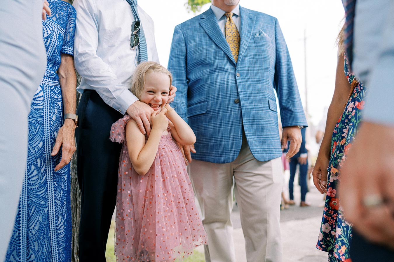 A young girl in a pink dress smiles while surrounded by well-dressed adults at an outdoor event.