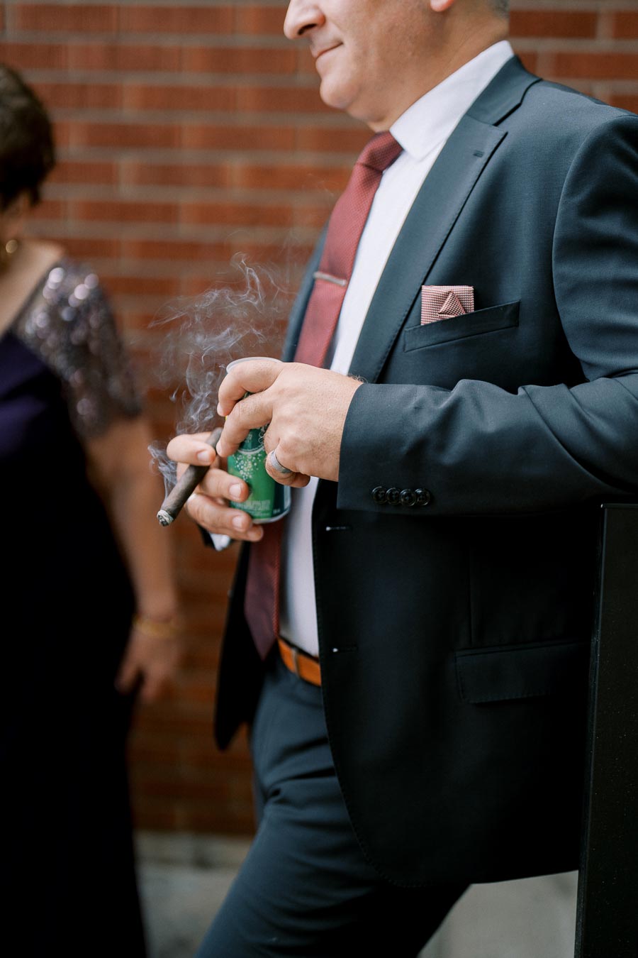 Elegant man in a suit leaning against a brick wall, holding a cigar and a can, exuding sophistication and style.