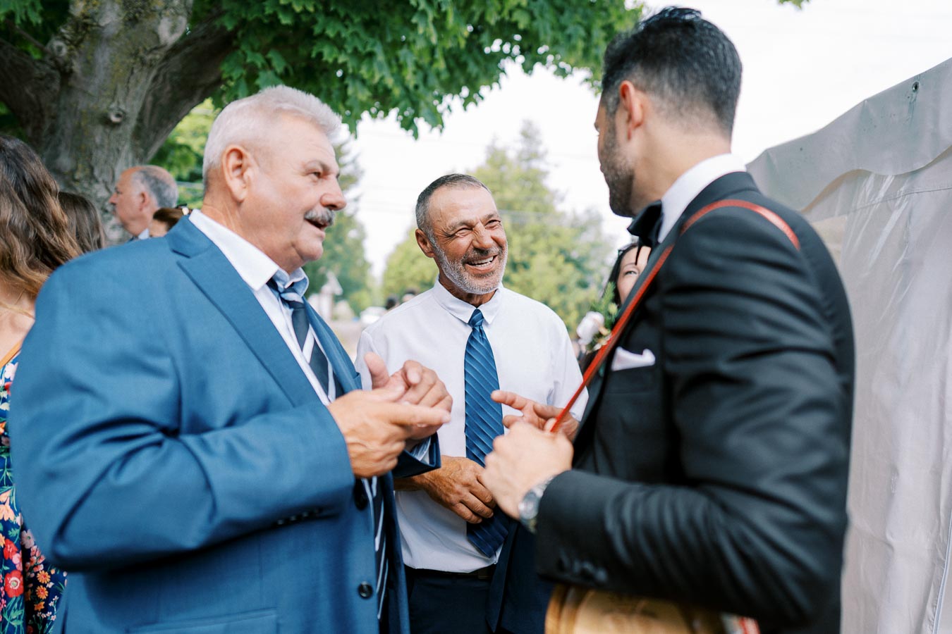 A group of well-dressed men engaged in conversation at an outdoor event, with greenery in the background.