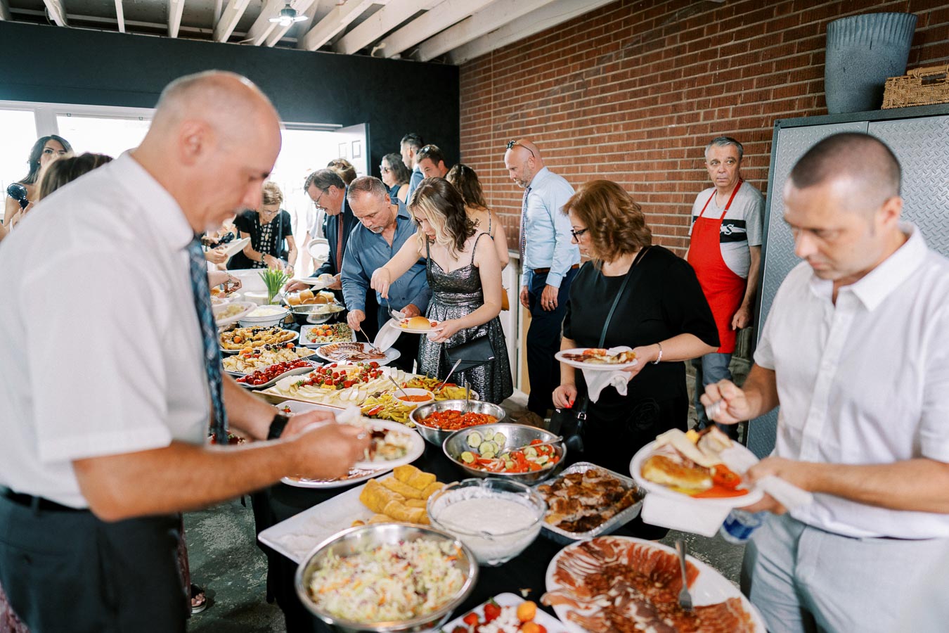 A group of people serving themselves from a buffet table filled with various dishes, including salads, meats, and appetizers, in a bright and spacious setting with a brick wall background.
